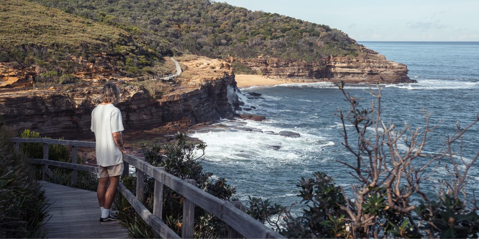 A man standing on a wooden boardwalk that hugs a coastline.
