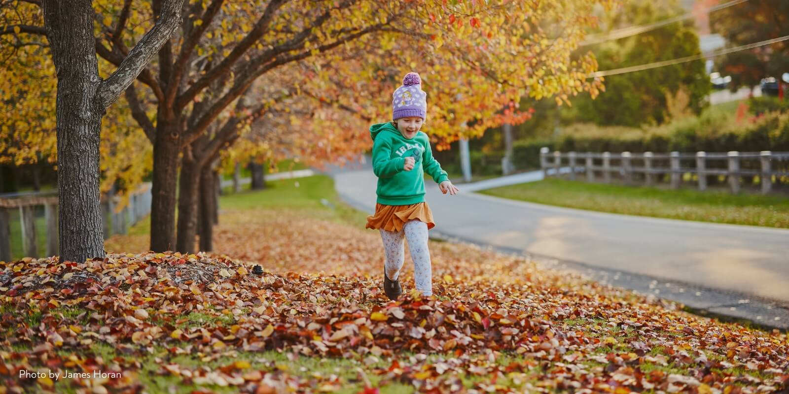 A girl wearing a bright green jumper and beanie running throughout a ground covered in fallen autumn leaves.