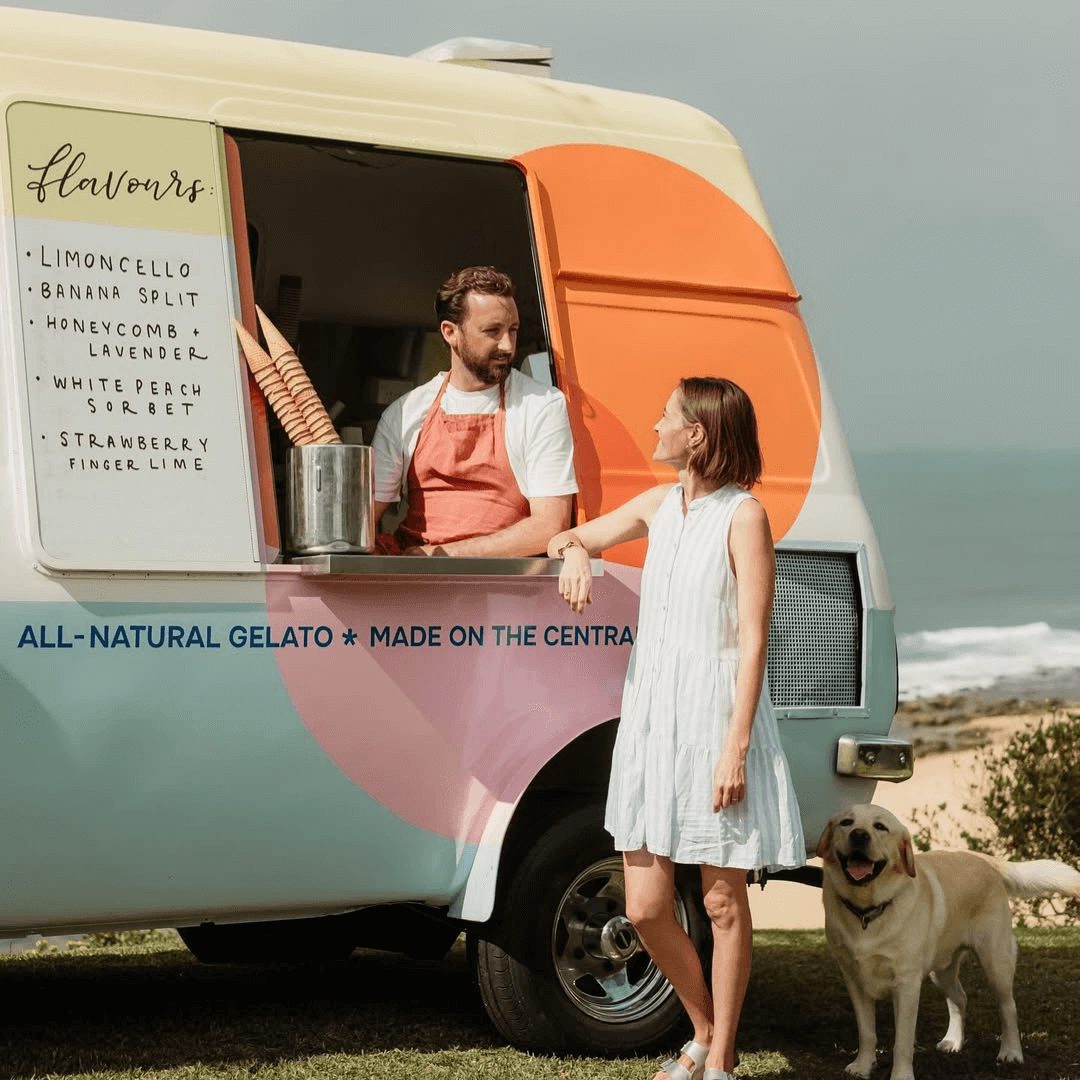 A lady hanging out beside a colourful van. A man is inside the van and a stack of icecream cones sit next to him