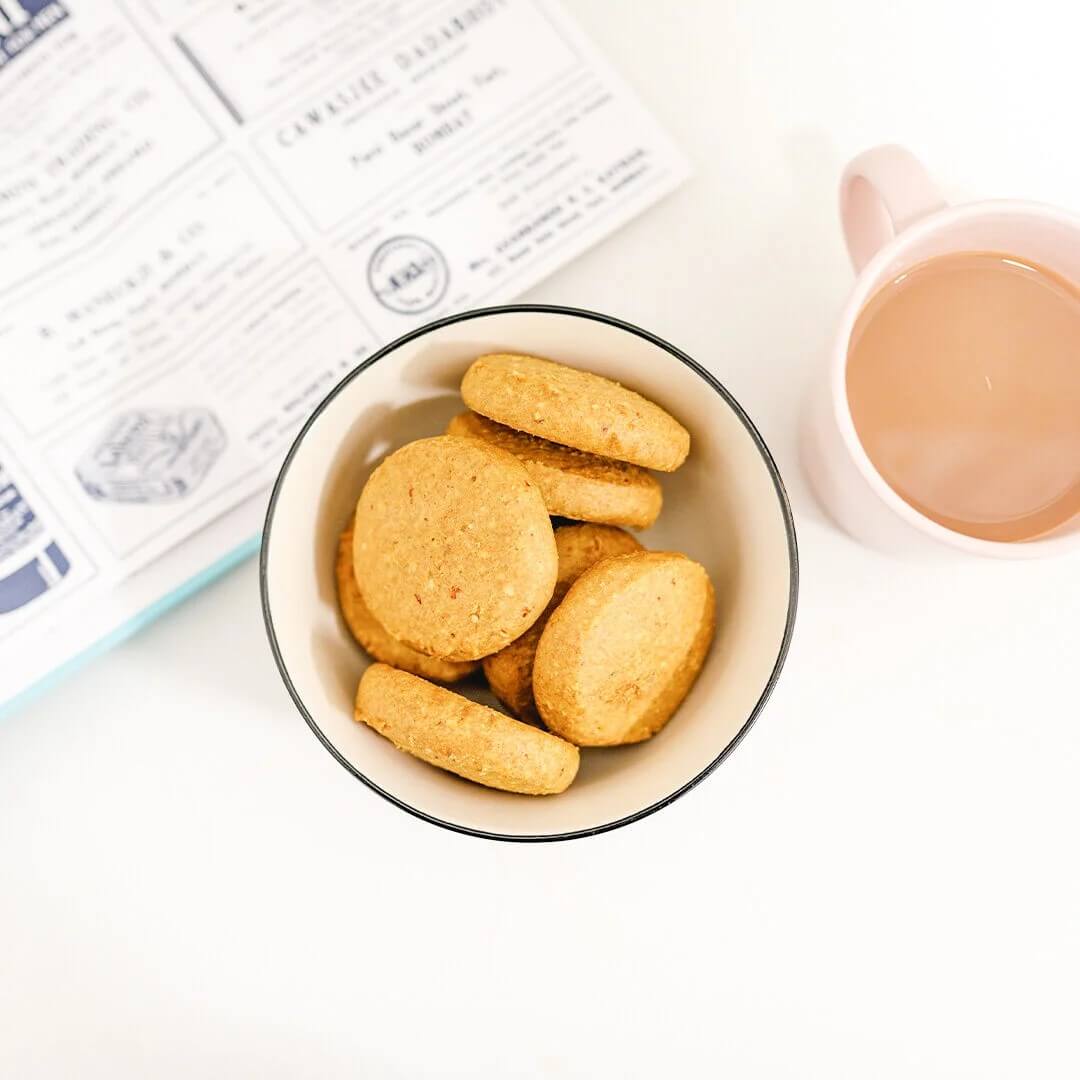 A cup filled with round brown biscuits and a tea in the background.