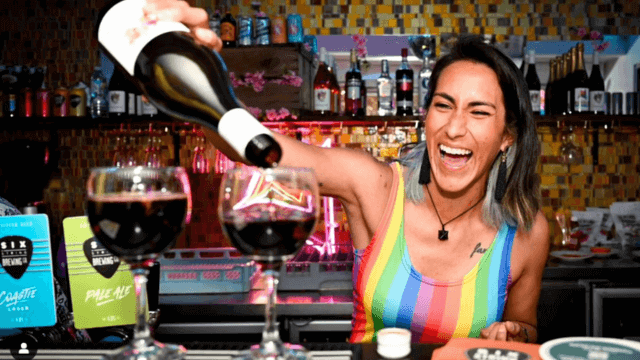 lady in rainbow top pouring 1 glasses of red wine.
