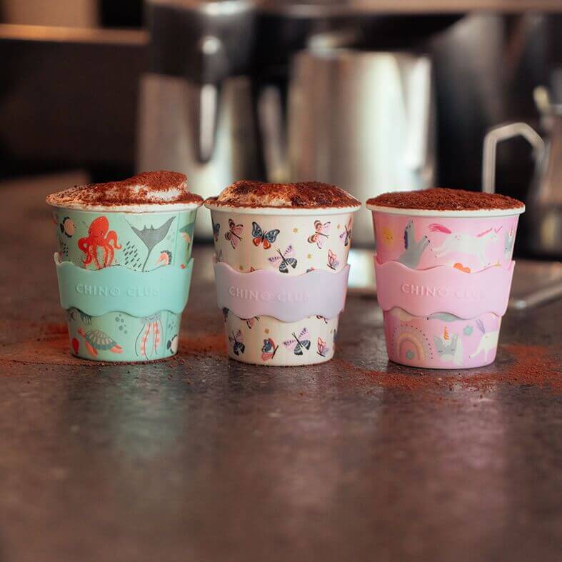 Three babyccino cups in green, cream and pink colours with milk froth and dusting of chocolate on top.
