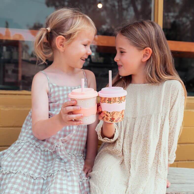 Two young girls sitting on a bench looking at each other doing a cheers with their babyccino cups with milk froth and dusting of chocolate on top. 