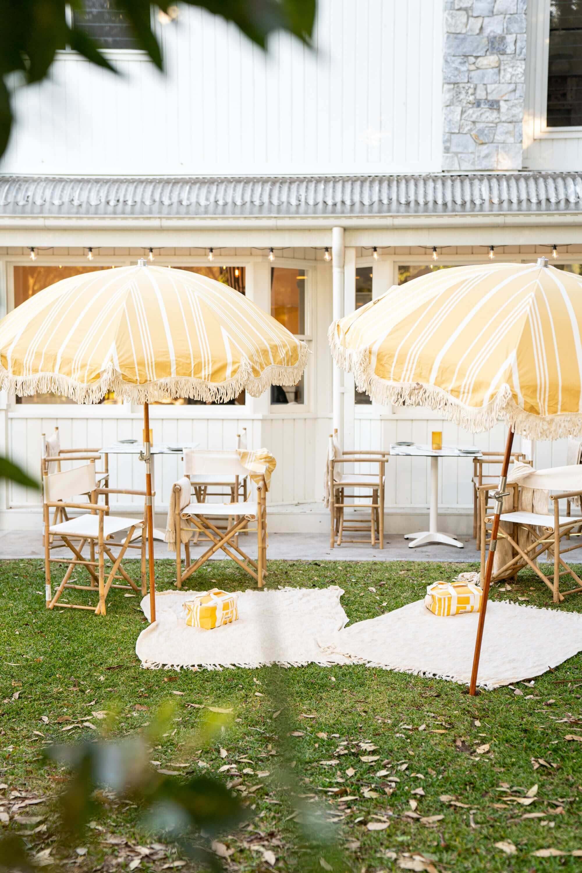 Outdoor picnic with yellow and white beach umbrellas with deck chairs, rugs and pillows on green grass in front of a white coastal styled restaurant.