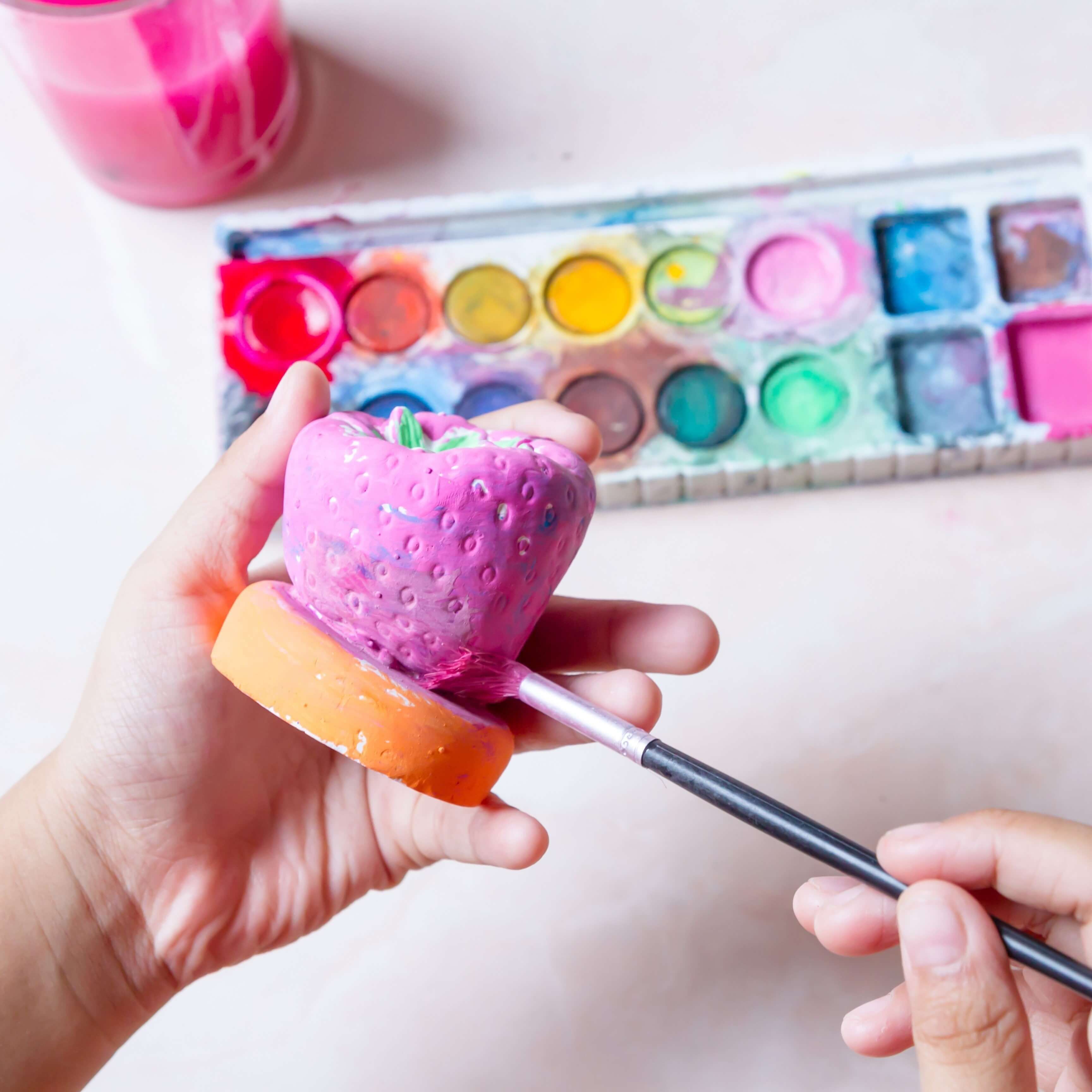 Bright and colourful paints with hands holding a plaster painting object painting it bright pink.