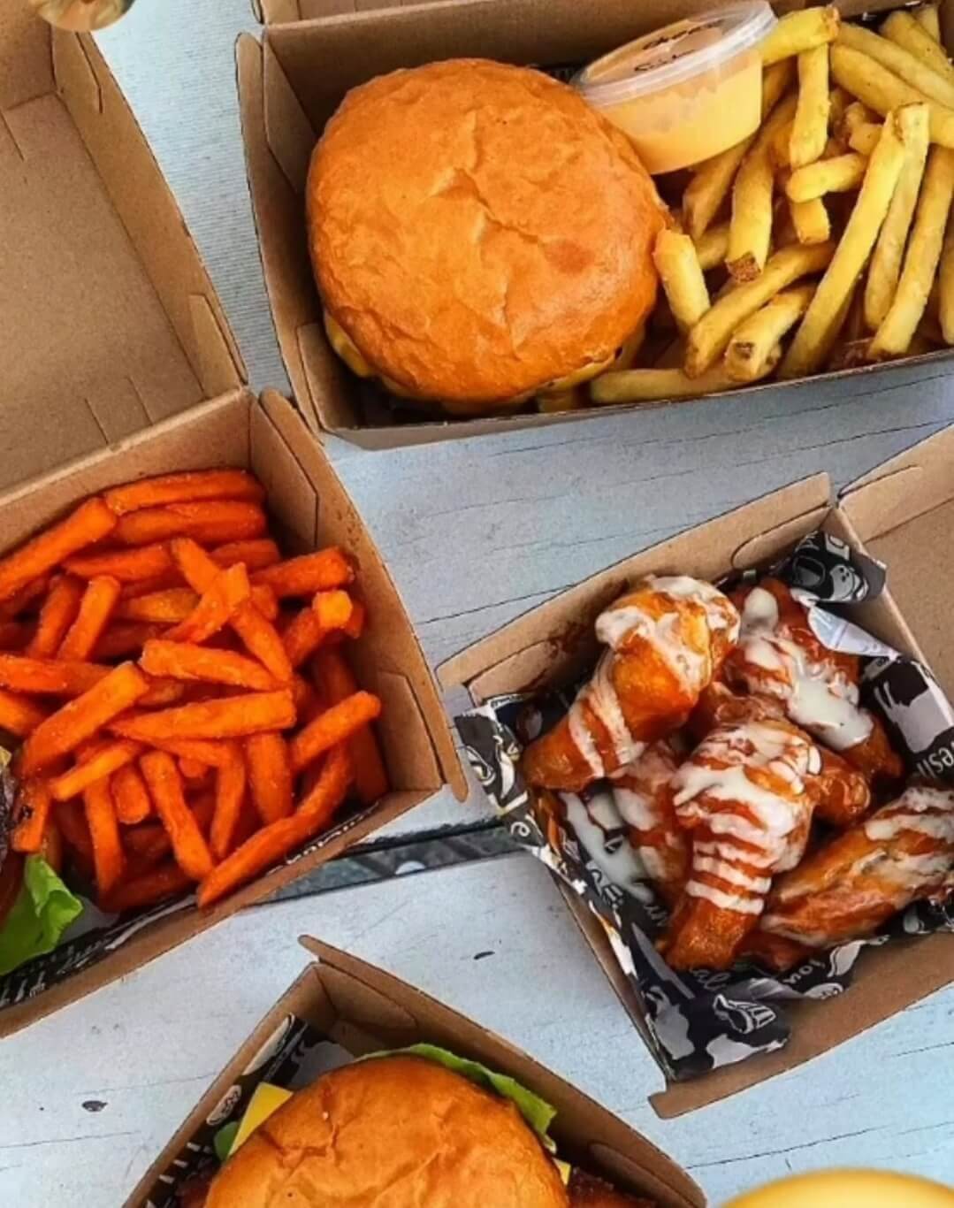 Overhead view of burgers, chips, sweet potato chips, chicken wings with a drizzle of sauce in cardboard recyclable boxes.