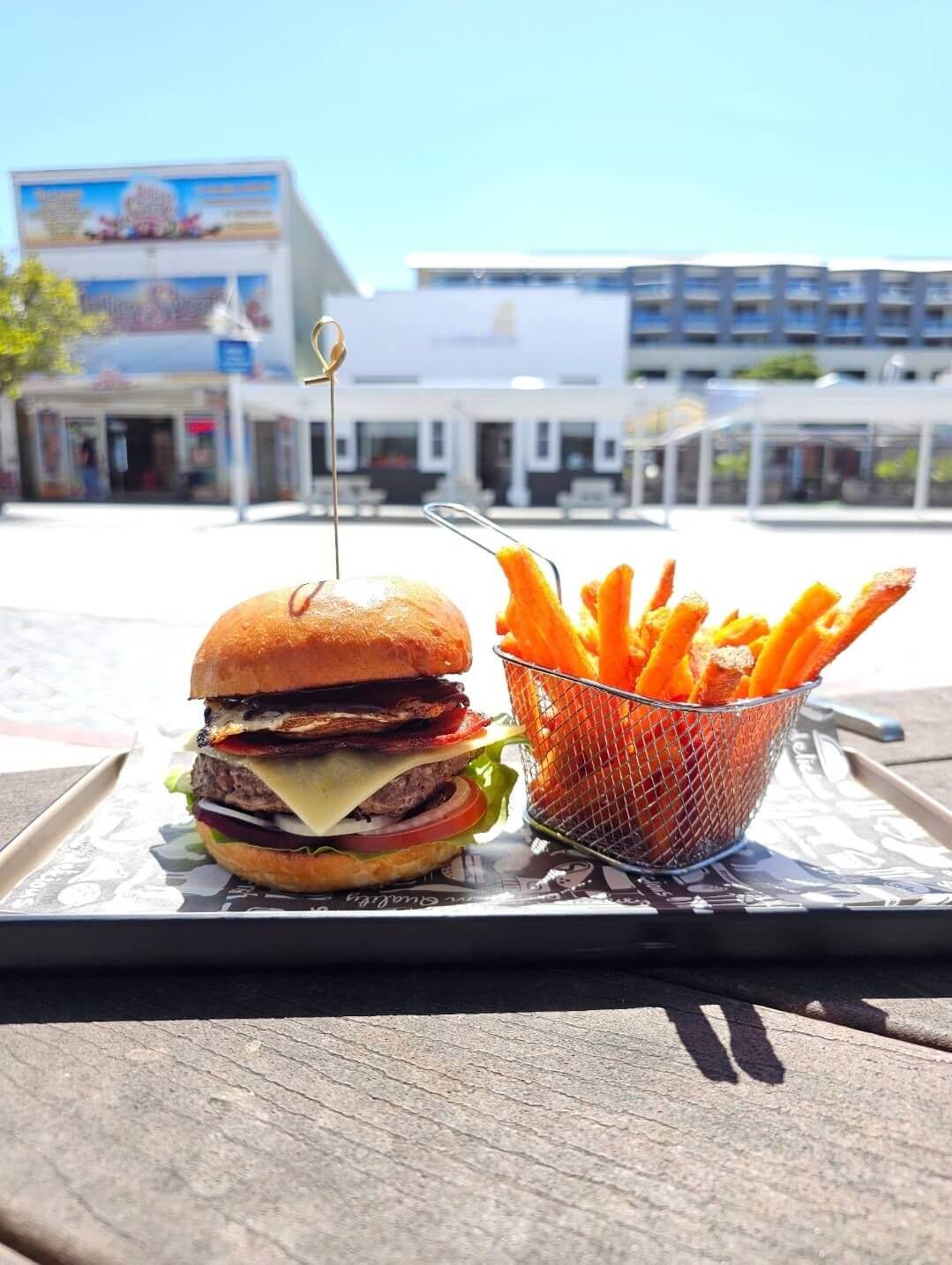Burgers and fries on a rectangle board in the sun with a row of shops in the distant background