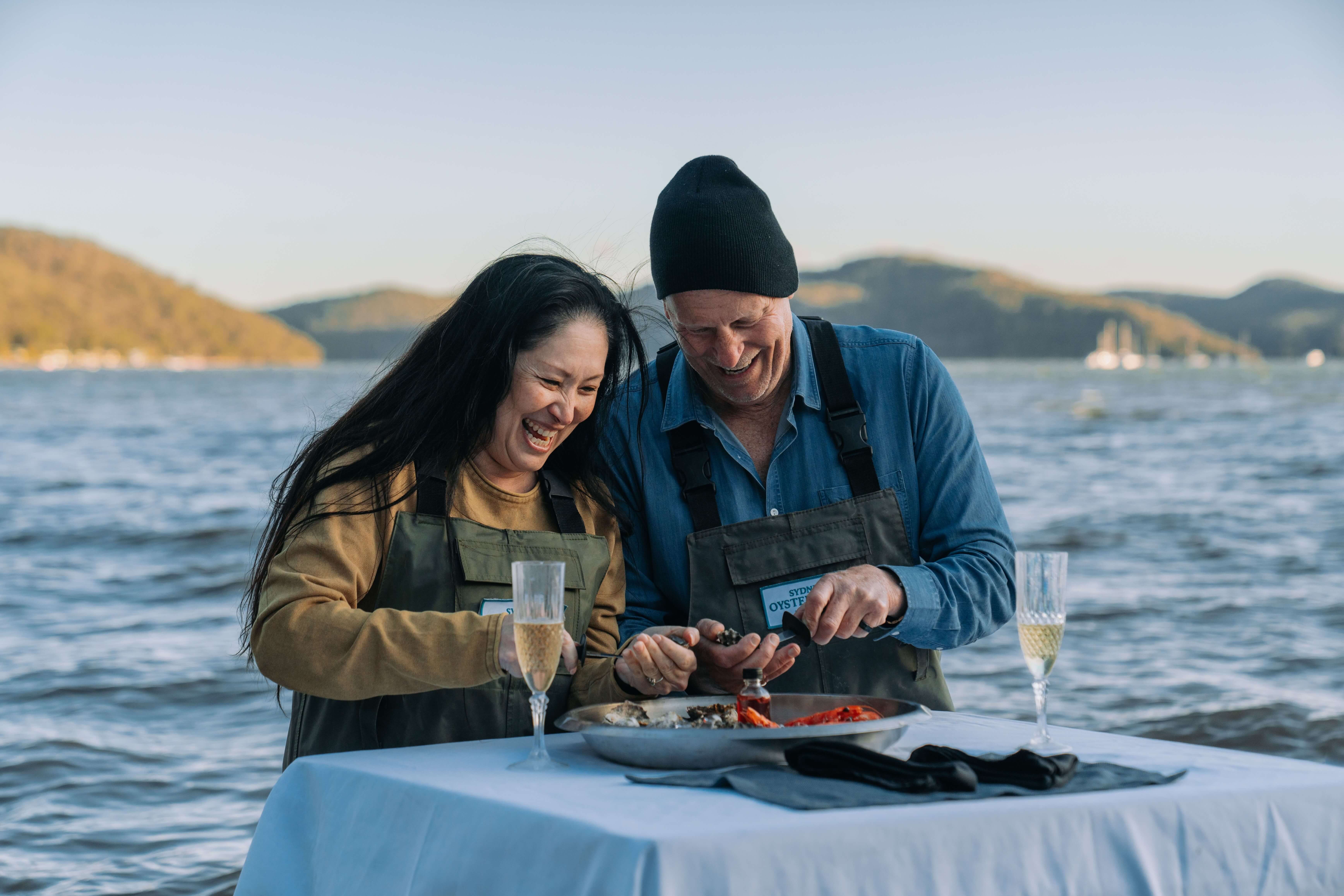 A couple wearing waders, shucking fresh oysters at a table that sits within the water