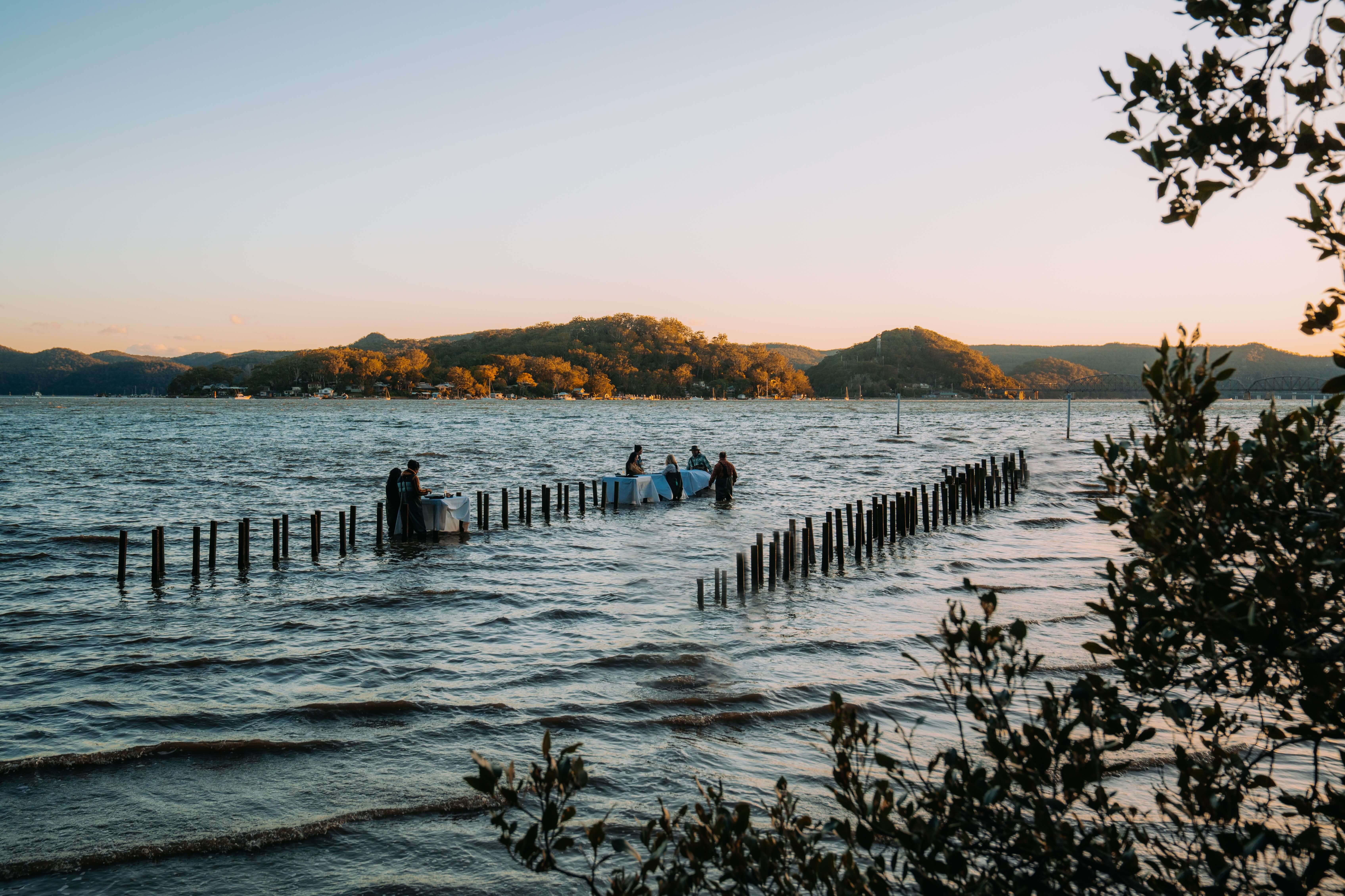 A river with two dining tables sitting in the water with people standing at them