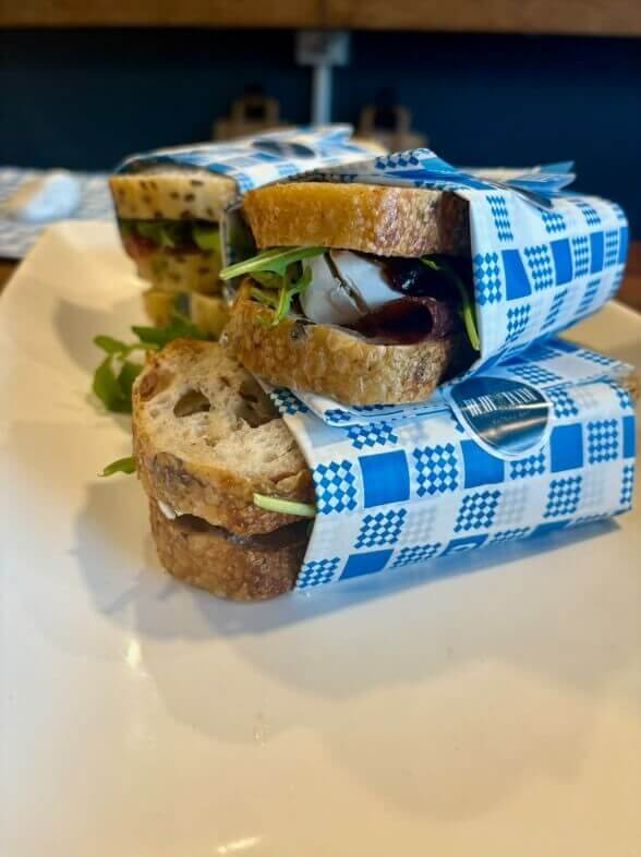 Three stacked handmade sandwiches wrapped in a blue tartan parchment paper.
