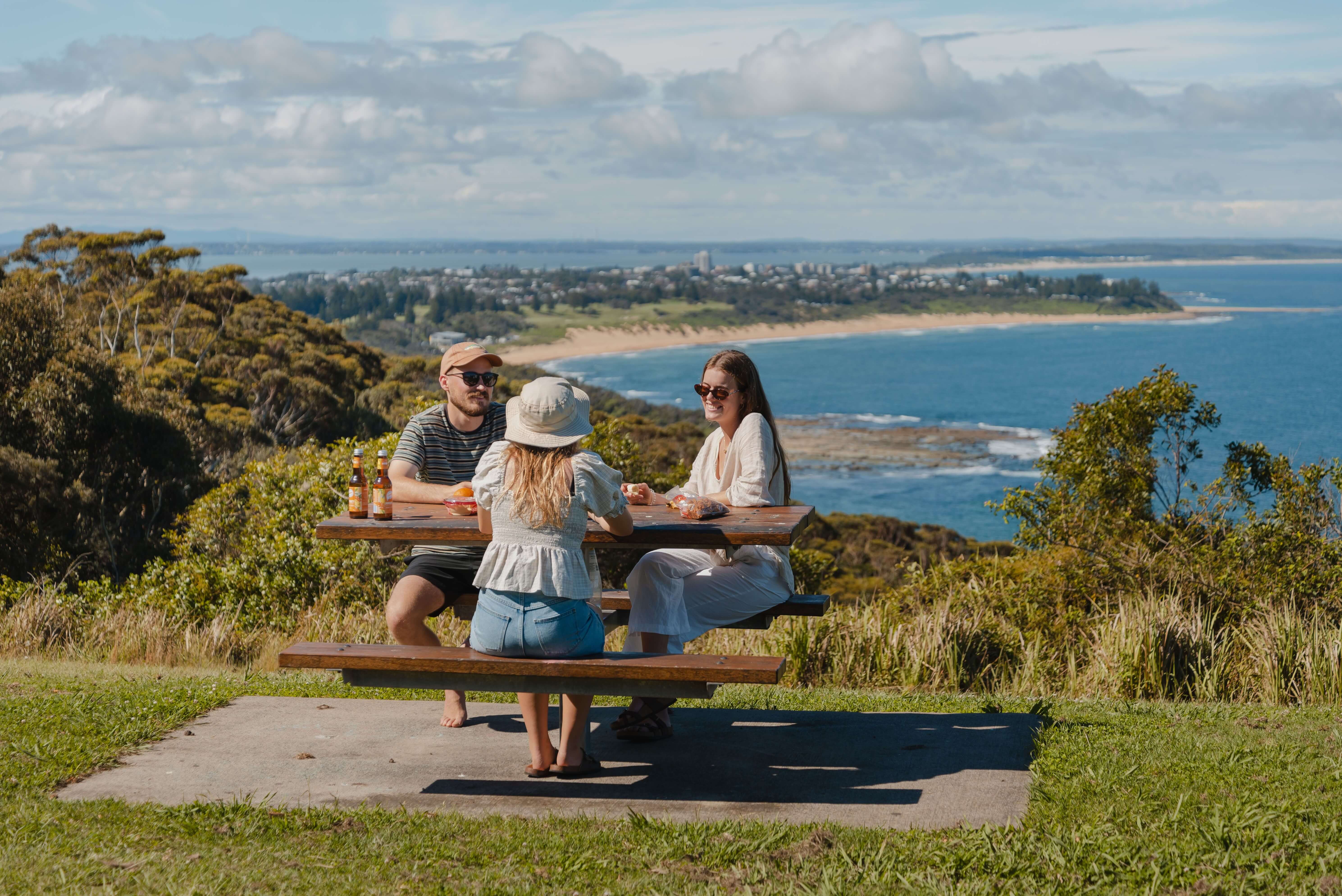 A group of friends enjoying lunch at a picnic table that overlooks a coastline