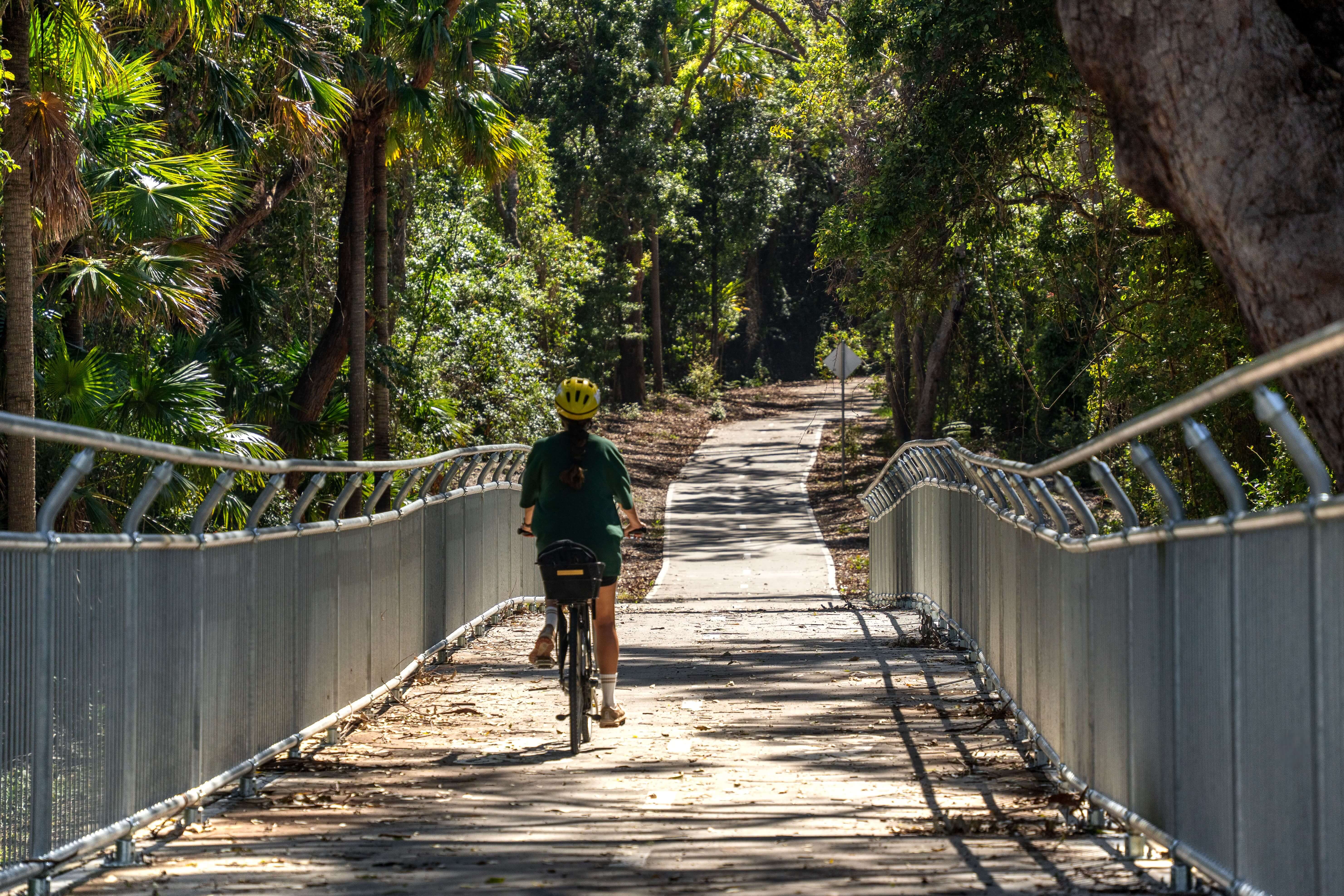 A lady with a yellow helmet and green jersey on a bicycle riding on a shared pathway through bushland
