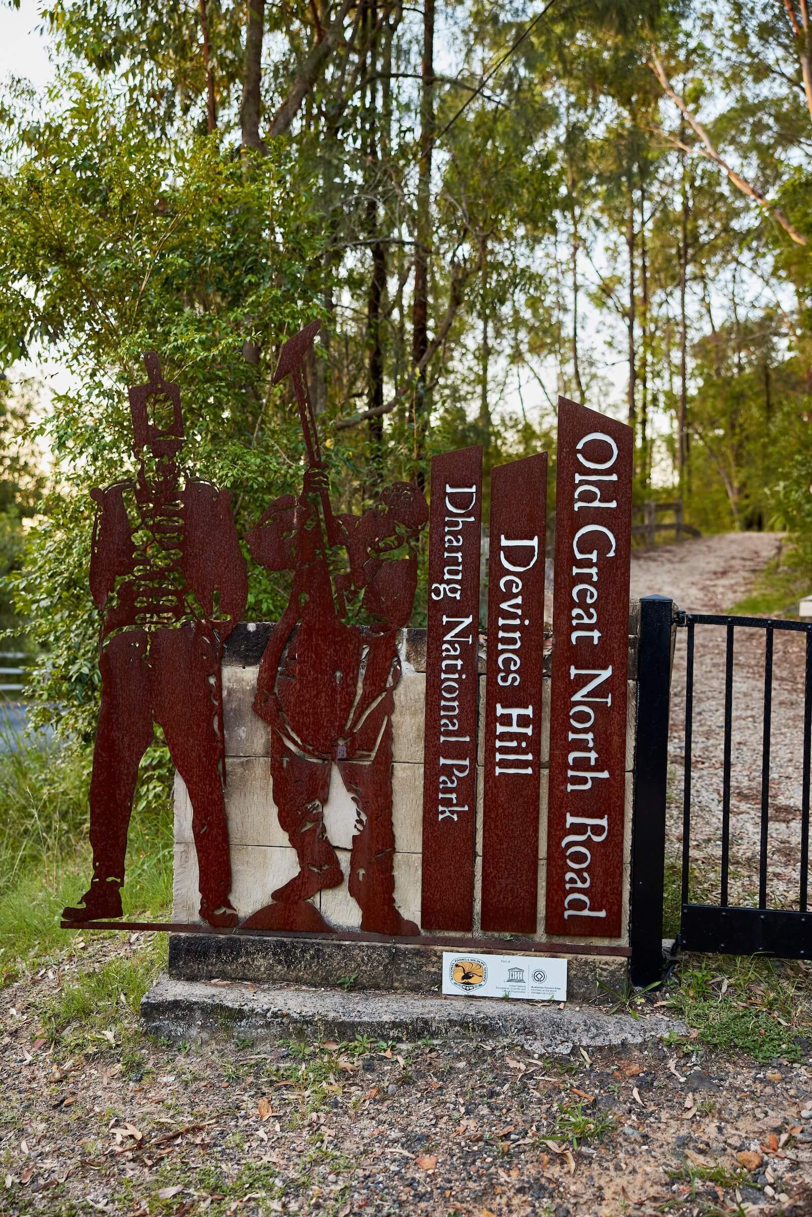 A dark brown-red cast iron gate and sign labelled Dharug National Park Devines Hill Old Great North Road
