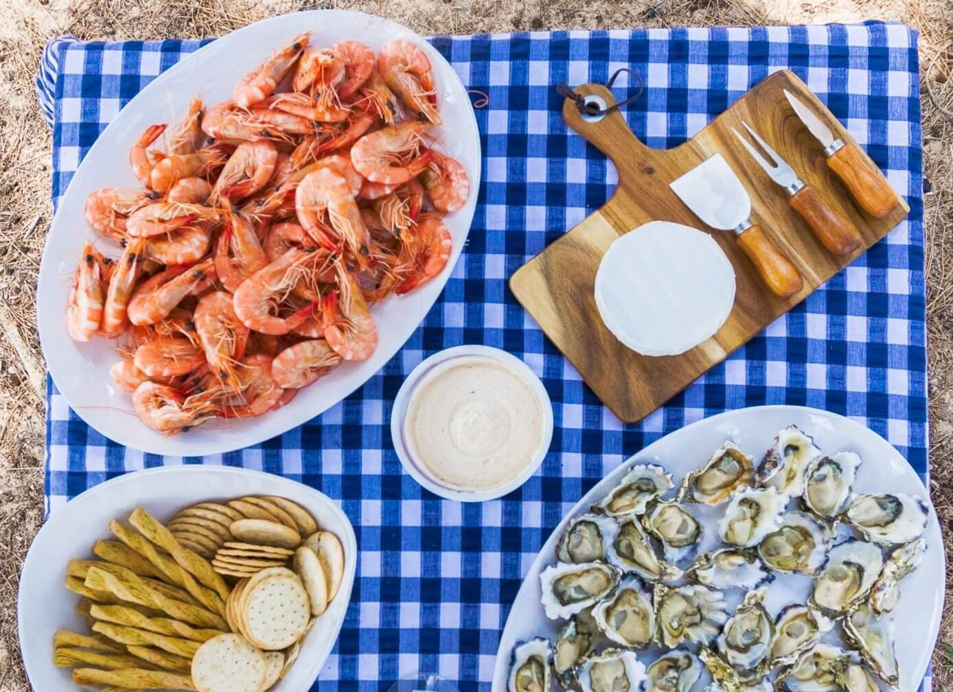Aerial view of a plate of cooked prawns, crackers, cheese platter and oysters on a blue and white checkered picnic blanket