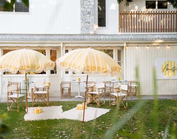 Outdoor picnic with yellow and white beach umbrellas with deck chairs, rugs and pillows on green grass in front of a white coastal styled restaurant.