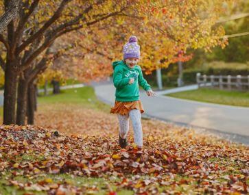 A girl wearing a bright green jumper and beanie running throughout a ground covered in fallen autumn leaves.