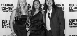 Three people in black and white stand against a premiere wall, smiling.